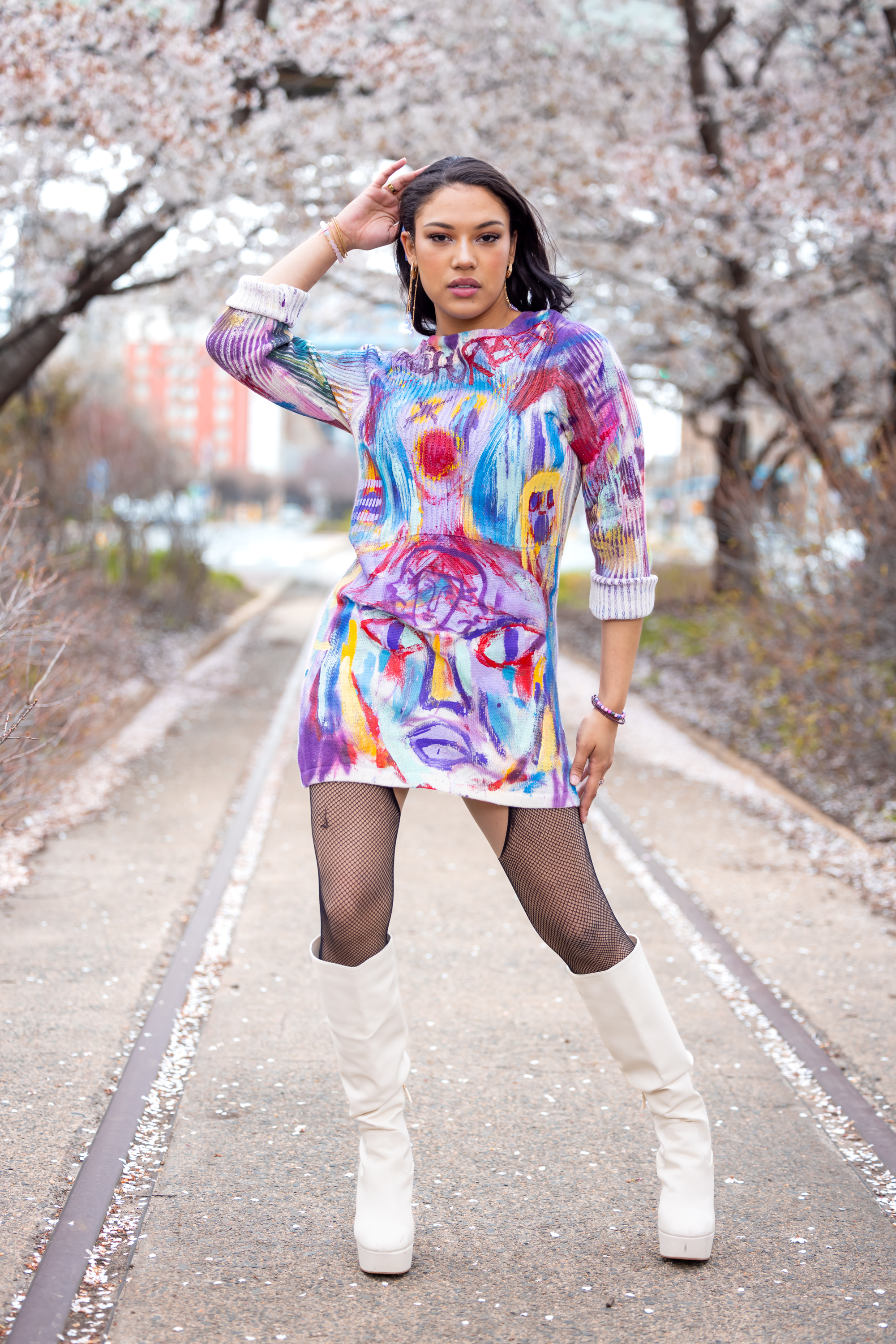 Model in a purple and multicolor hand-painted sweater dress with face artwork among cherry blossoms and white platform boots