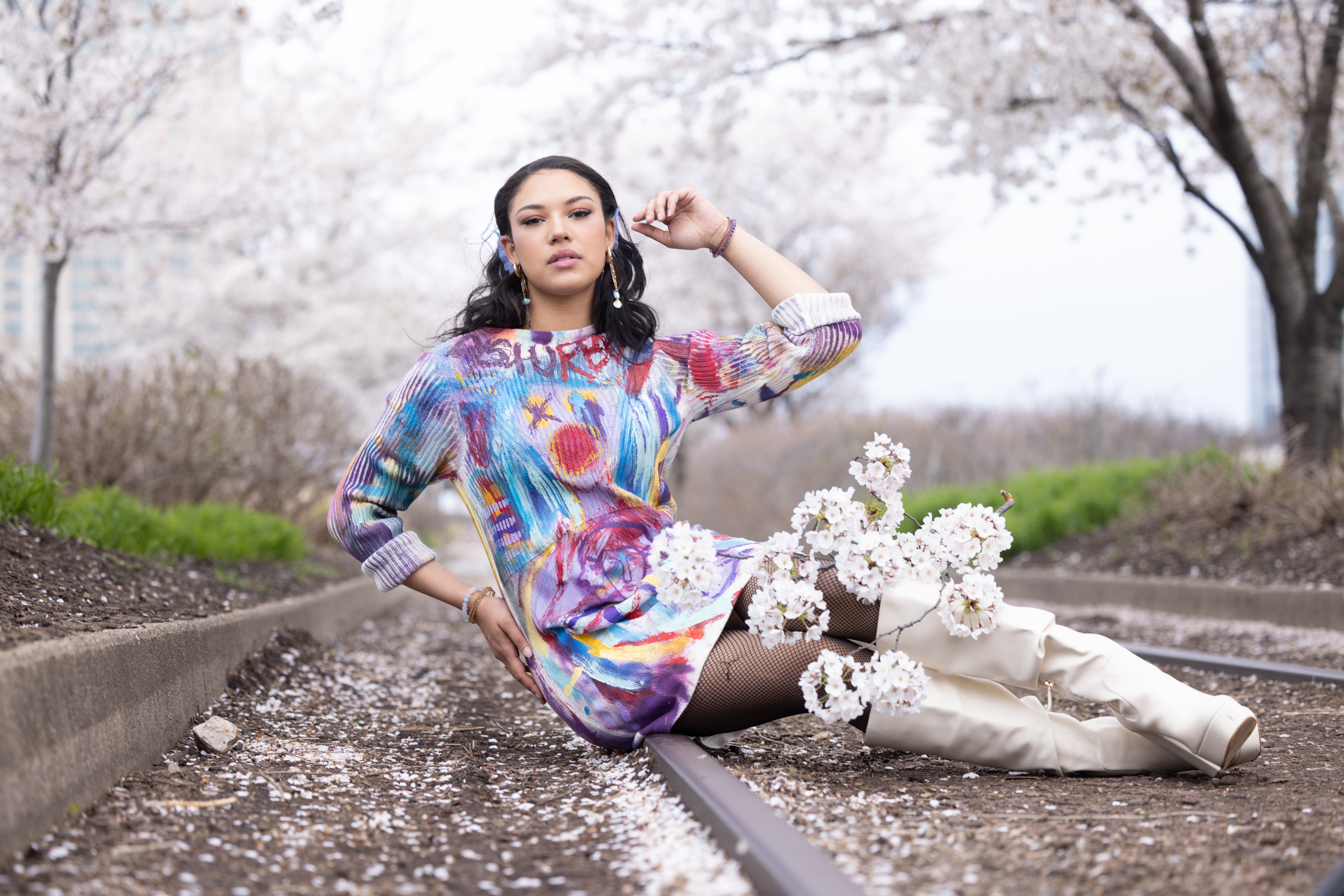 Model seated on cherry blossom railroad tracks in a multicolor hand-painted sweater dress with white boots