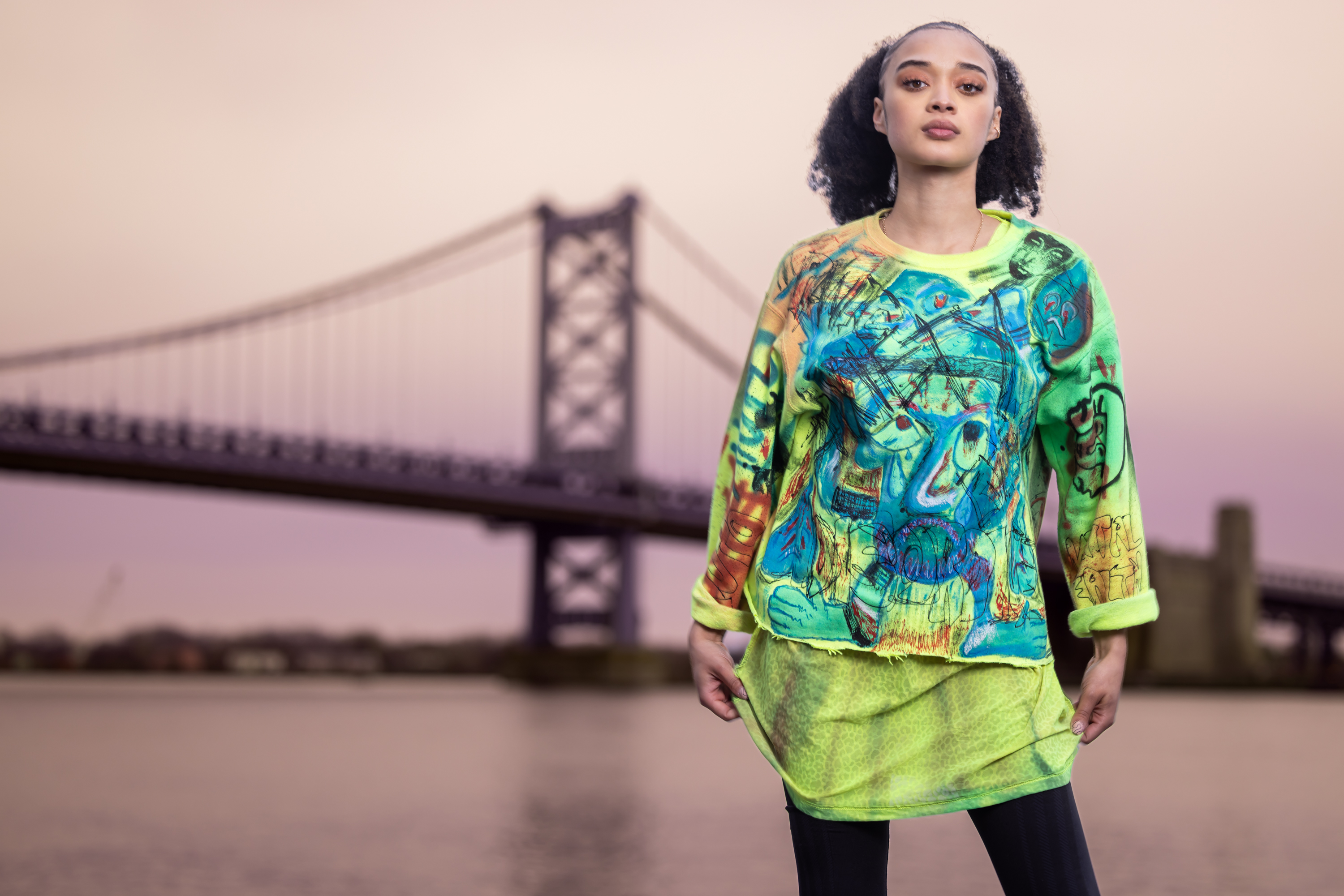Model in a green and yellow hand-painted graffiti sweatshirt with the Ben Franklin Bridge at sunset