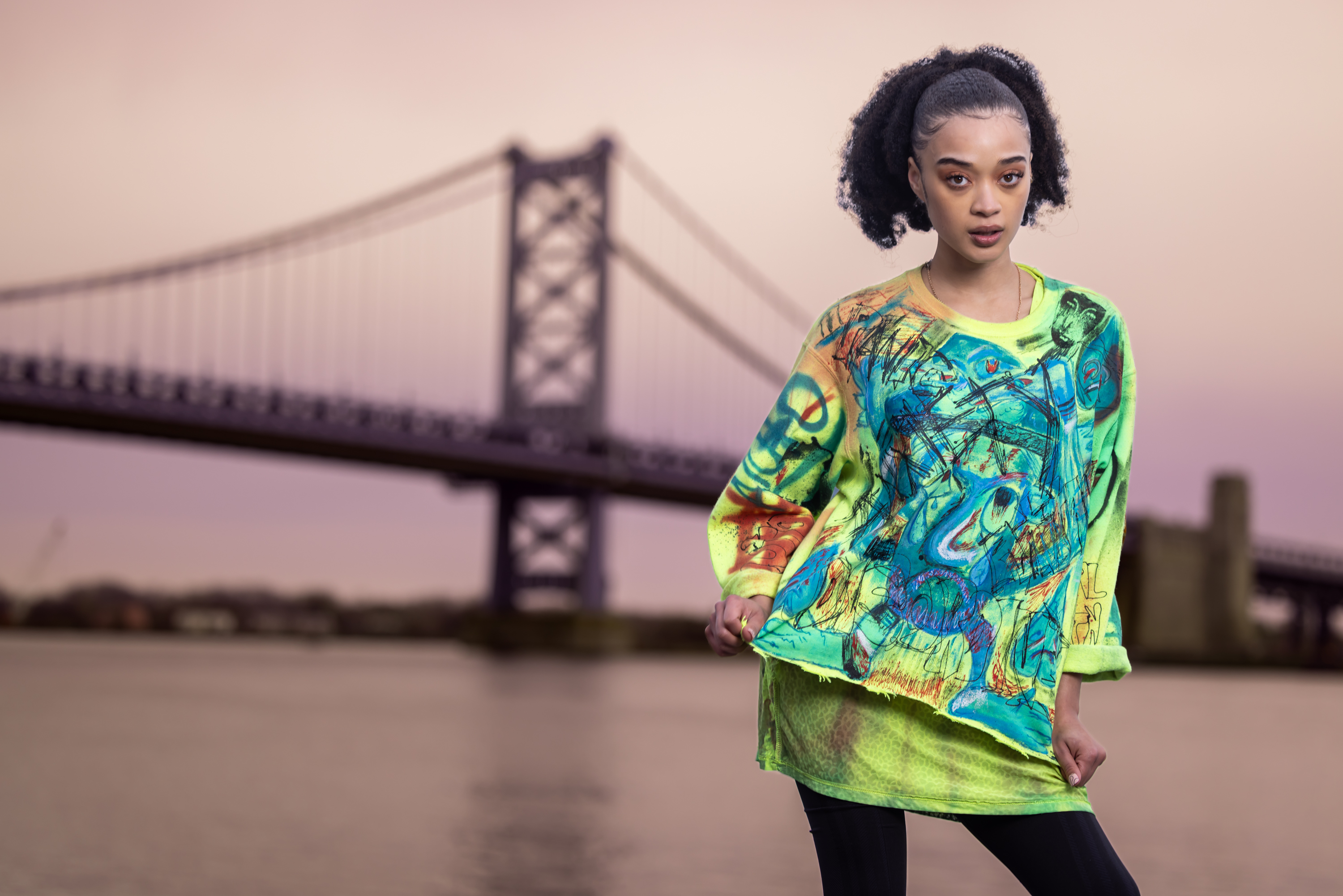 Model in a green and yellow hand-painted graffiti sweatshirt by the Ben Franklin Bridge at sunset