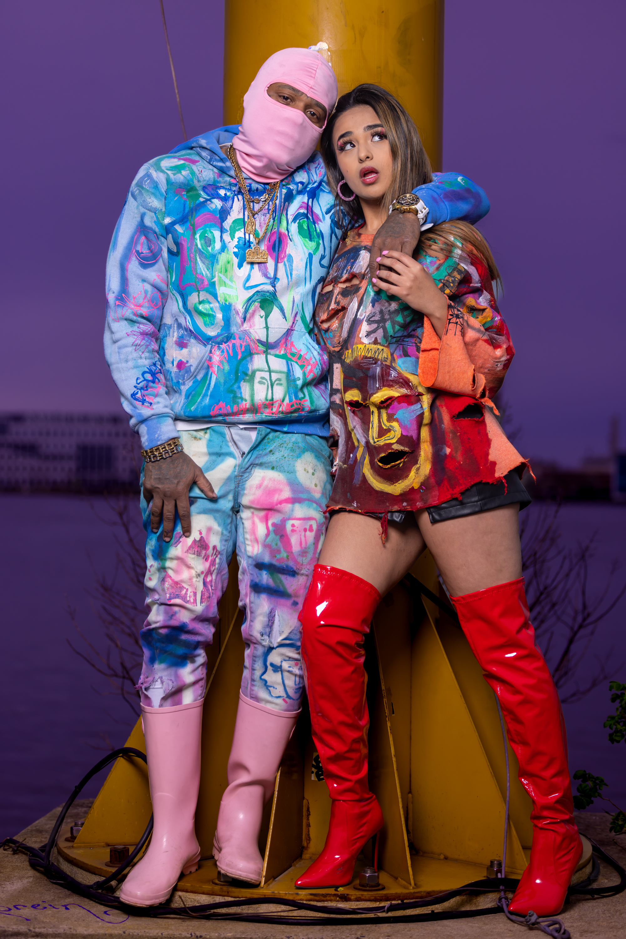 Franco in a pink mask and blue hand-painted tracksuit with a model in a red face-art dress and red boots by the yellow piling at dusk