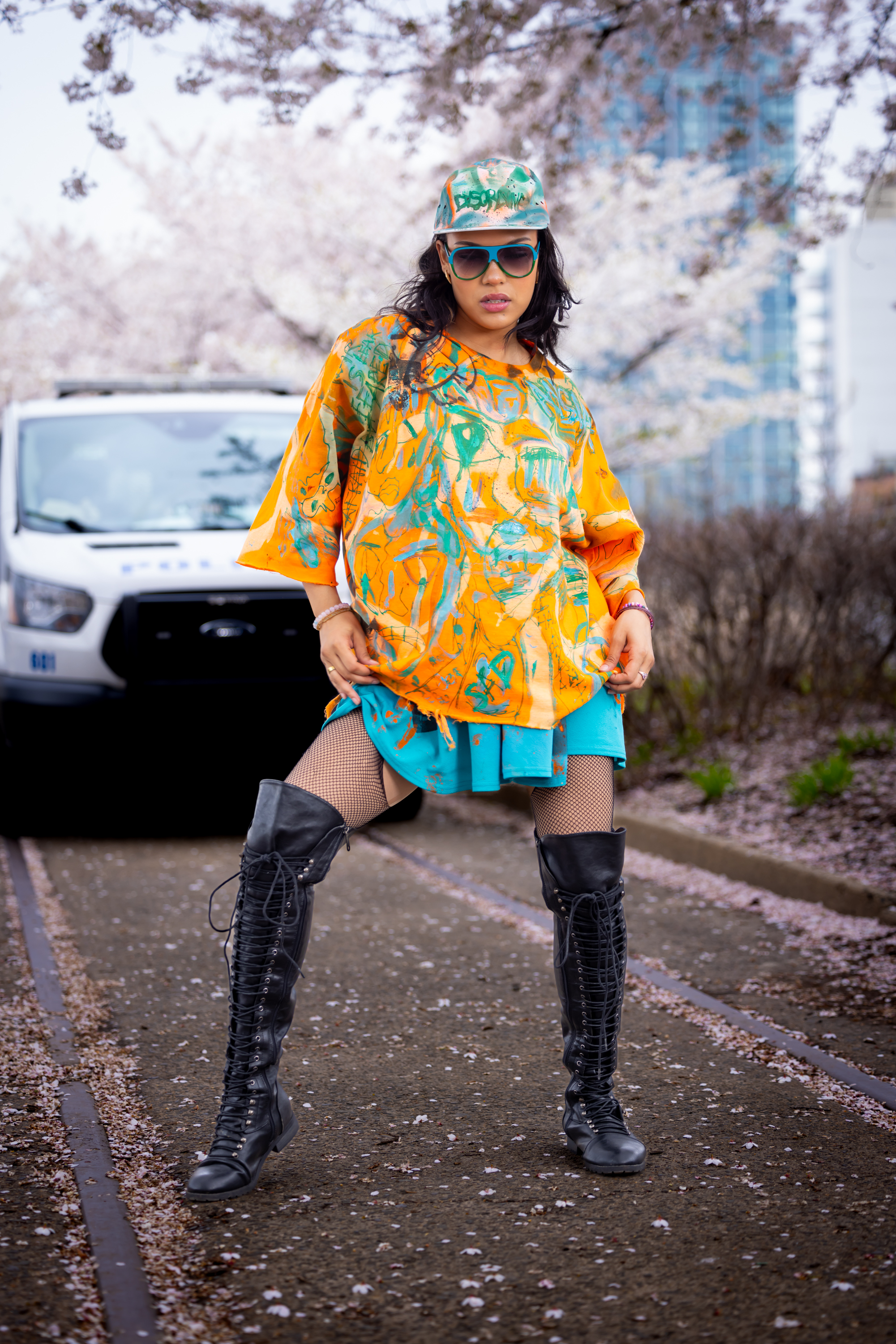 Model in an orange and teal hand-painted DiSoRdInE tee, cap, and black boots on a cherry blossom path