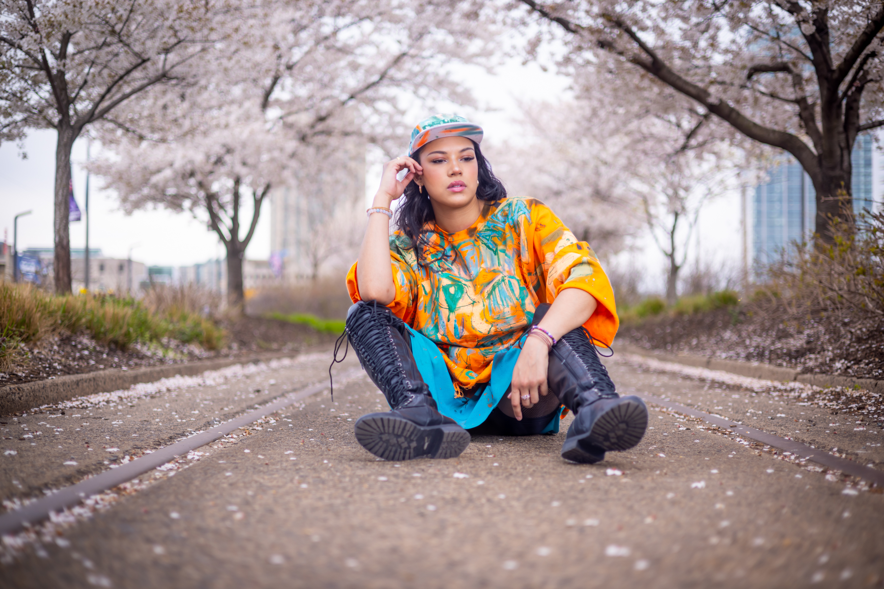 Model seated on cherry blossom railroad tracks in an orange and teal hand-painted DiSoRdInE tee and cap