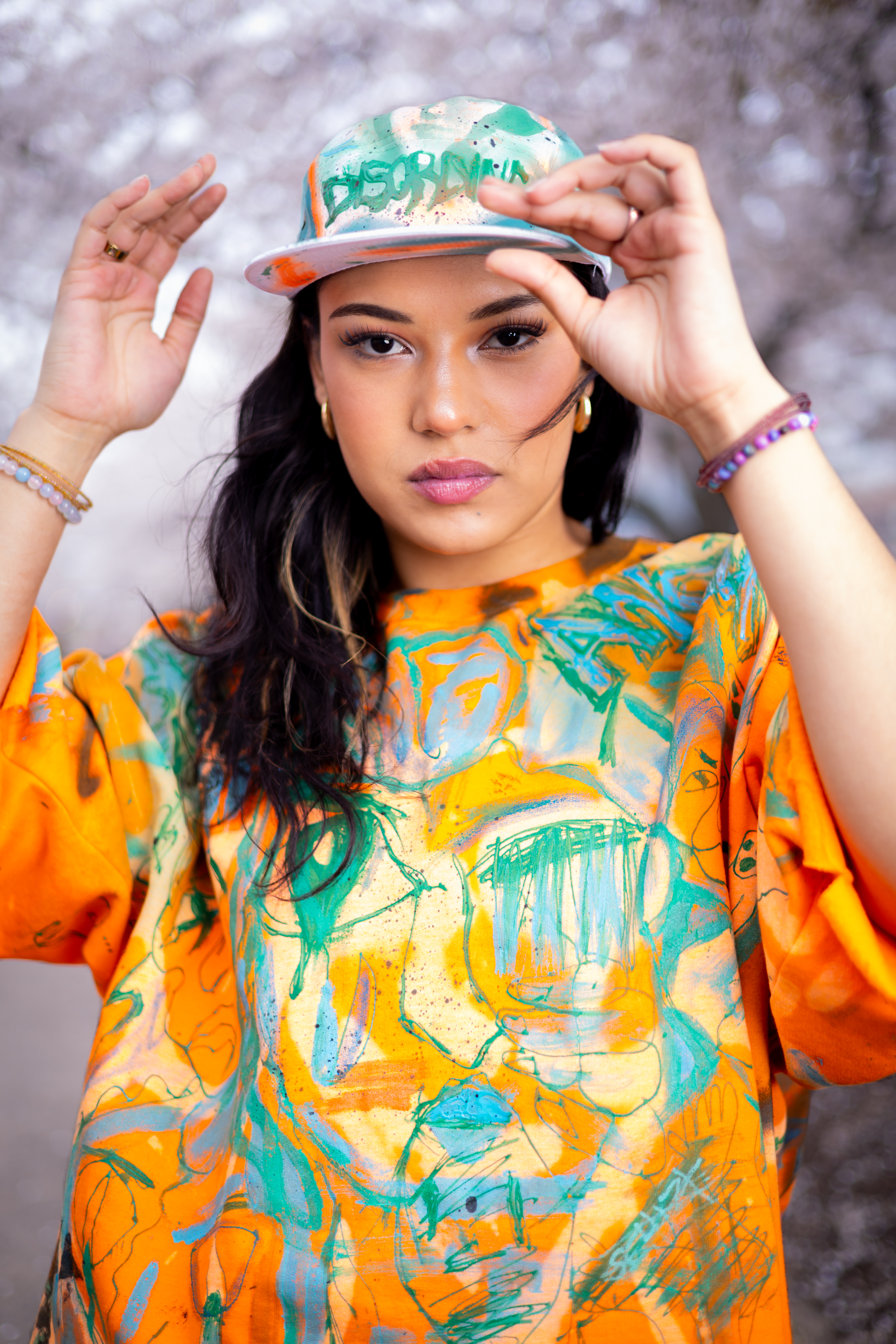 Close-up of a model in an orange and teal hand-painted DiSoRdInE tee and cap among cherry blossoms